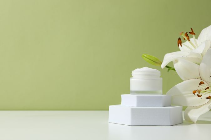 Jar with cream and lilies on green background, space for text