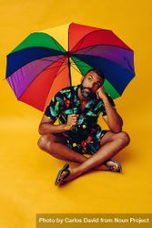 Happy Black male thinking about something while sitting under colorful umbrella 0vZYB0