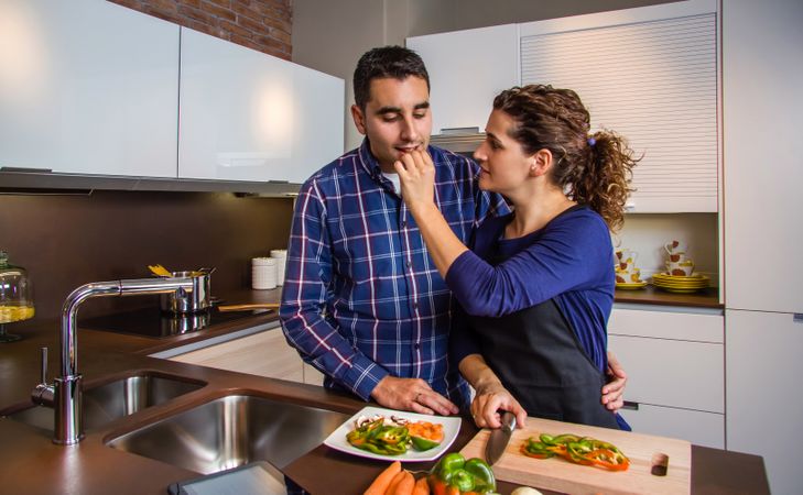 Couple prepping food in kitchen while wife feed vegetable to husband
