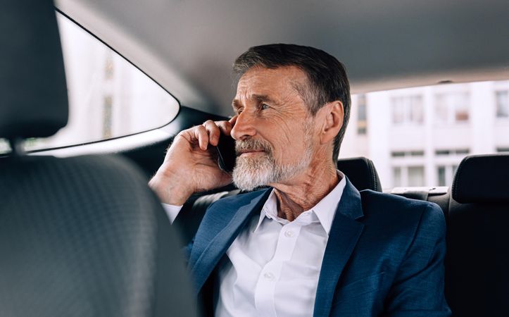 Mature man with grey beard taking a call in backseat of car