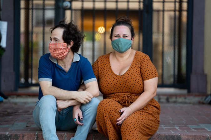 Wife and husband holding hands sitting together on stoop in front of apartment wearing masks