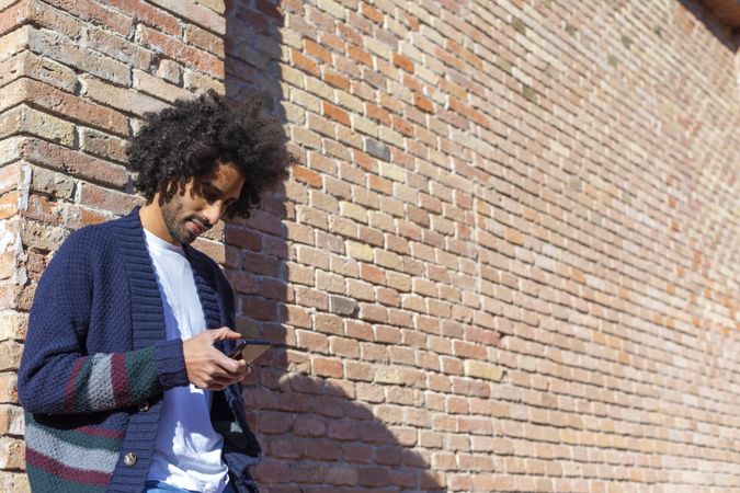Curly haired man using his smartphone while leaning on a brick wall outdoors on sunny day