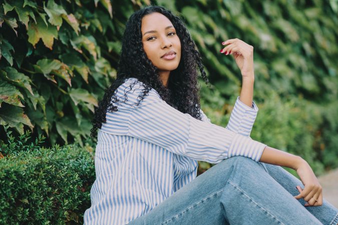 A Stylish Young Woman Sitting Comfortably Against a Backdrop of Lush Greenery in Casual Attire