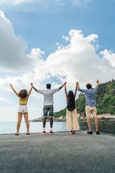 Vertical shot of diverse friends holding hands with arms raised, celebrating on a pier by the sea.