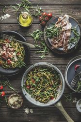 Italian food background with green cooked pasta in cooking pan with parma ham, arugula, tomatoes and cheese on rustic wooden kitchen table.
