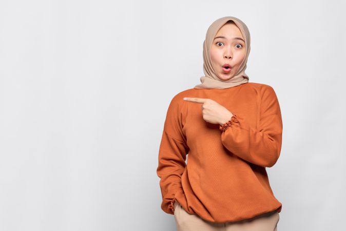 Surprised Muslim woman in headscarf and orange sweater pointing to her side