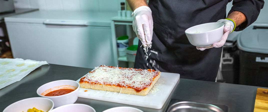 Banner of anonymous chef sprinkling grated cheese over a tomato sauced pizza in restaurant kitchen