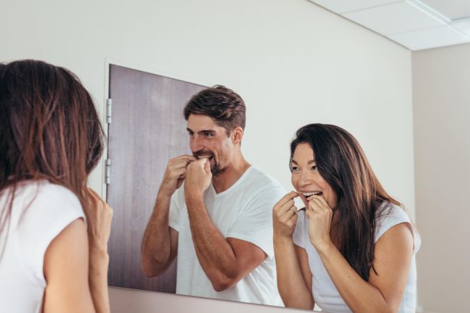 Couple looking in the mirror and smiling, using dental floss to clean their teeth