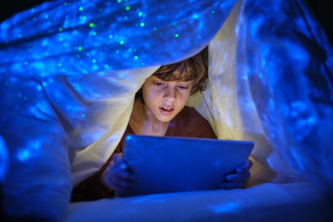 Boy playing game on tablet under blanket in evening