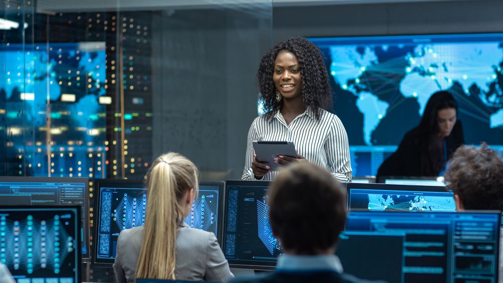 Chief Project Engineer Consults Tablet Computer and Holds Briefing for a Team of Scientists that are Building Machine Learning System.