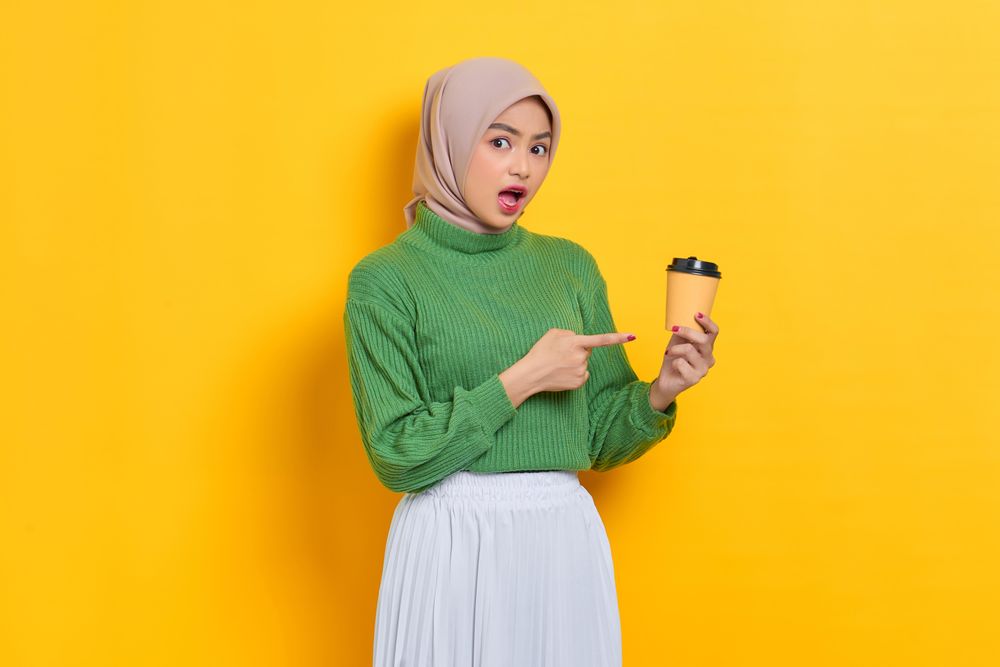 Surprised woman in headscarf pointing to to go coffee cup