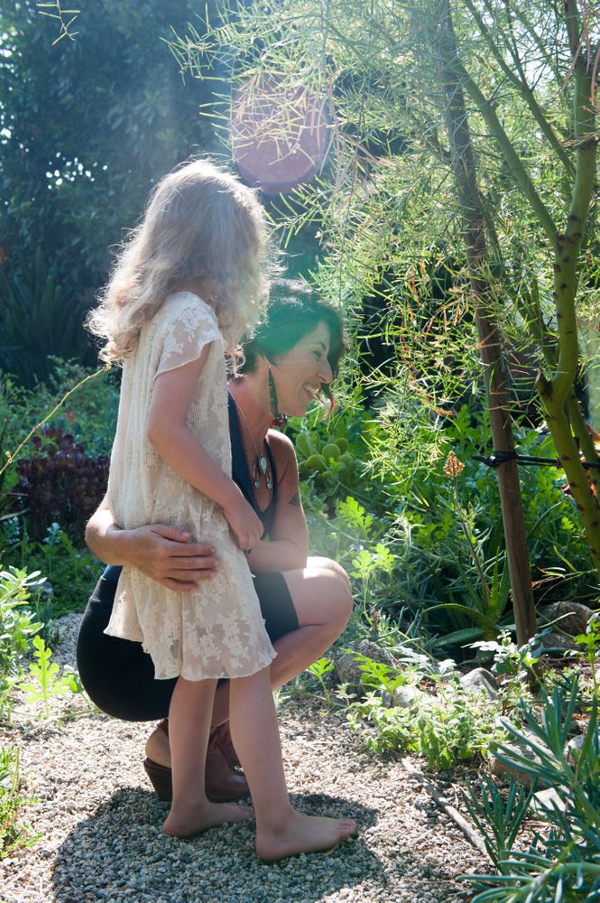 Smiling mother puts arm around  daughter on rock path exploring plants outside with lens flare