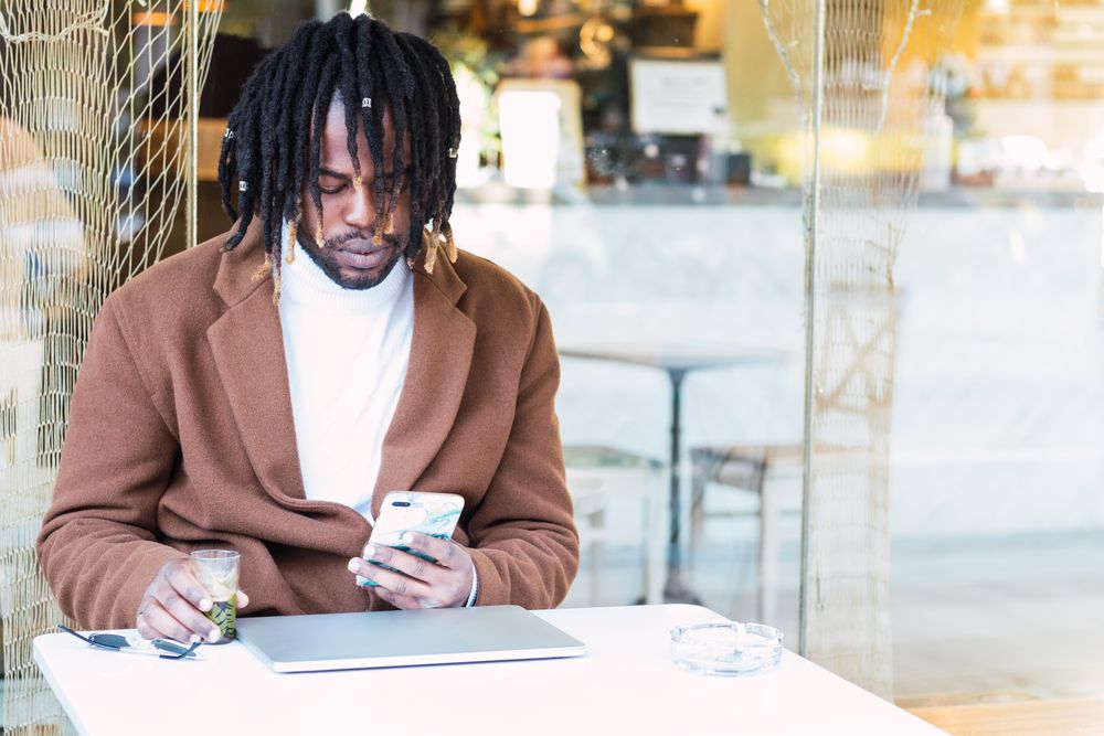 Black man with laptop browsing on mobile phone on restaurant terrace