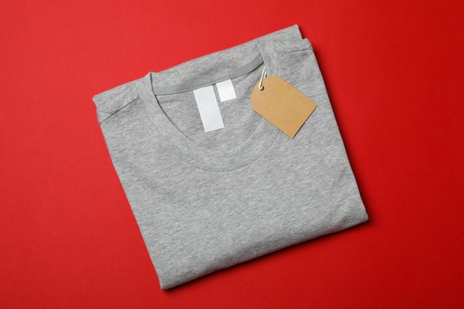 Folded blank grey t-shirt with tag on red background, space for text