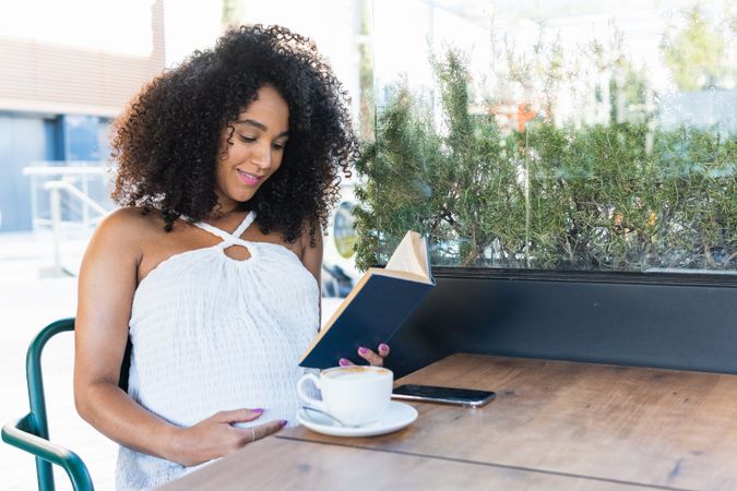 Pregnant Black woman reading book and drinking coffee