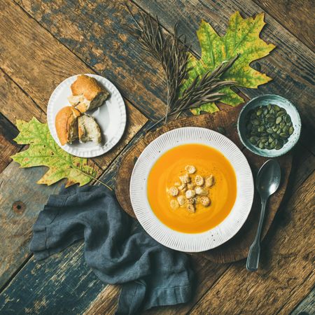 Pumpkin soup with croutons, bread, seeds, spoon on wooden table with leaves, square crop
