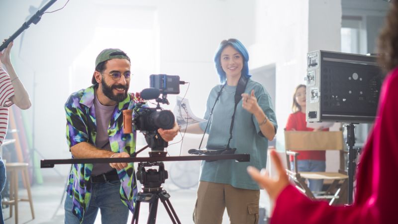 Young Male And Female Filmmakers Collaborate On A Film Set, Adjusting Camera Equipment With Enthusiasm, Surrounded By Diverse Crew Members In A Bright, Modern Studio Environment.