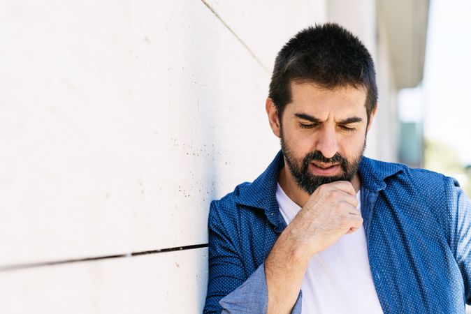 Latino man in denim standing outside holding beard deep in thought