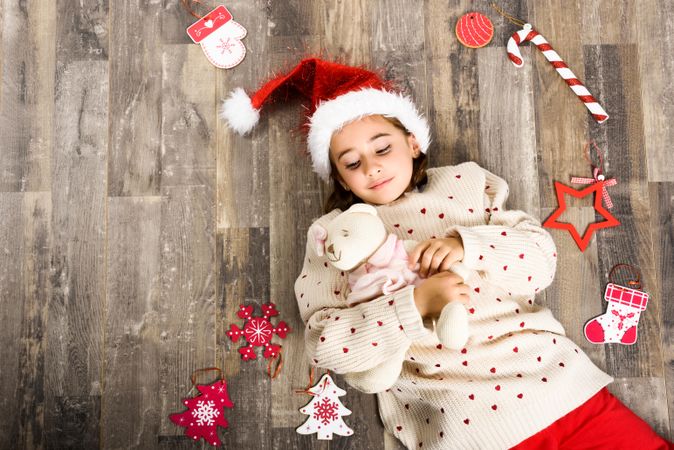 Girl in Santa hat lying on wooden floor surrounded by presents
