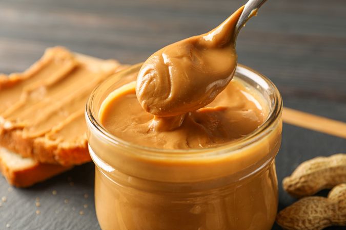 Glass jar with peanut butter and spoon, peanut, peanut butter sandwich and cutting board on wooden background, space for text and closeup