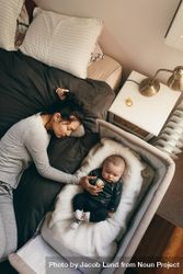 Top view of a young mother sleeping on bed with her baby sleeping on a bedside crib 0WXar5