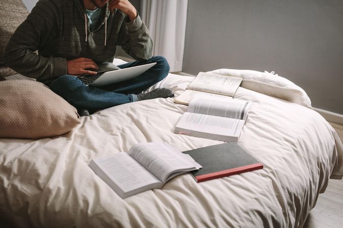 Cropped shot of student sitting on bed with books and laptop
