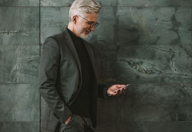 Mature businessman leaning to a wall and looking at cellphone