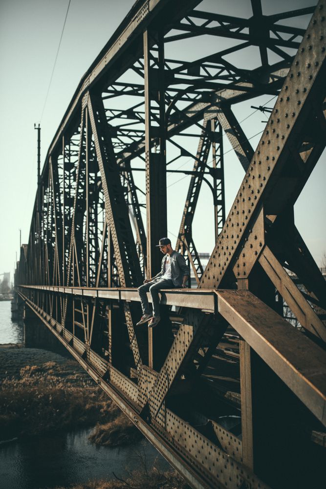 Man in blue denim jacket sitting on rusty brown iron bridge