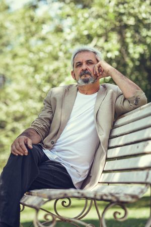 Portrait of a grey haired man, sitting on bench in a park deep in thought, vertical