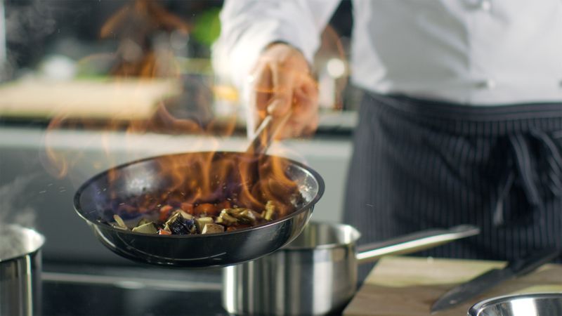 Professional Chef Cooks Flambe Style. He Prepares Dish in a Pan with Open Flames. He Works in a Modern Kitchen with Different Ingredients Lying Around.