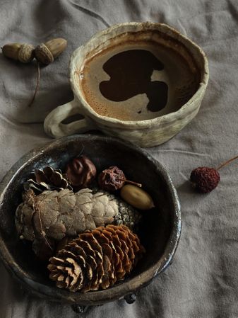 Dark coffee in ceramic mug on bed with bowl of pine cones with grey sheets