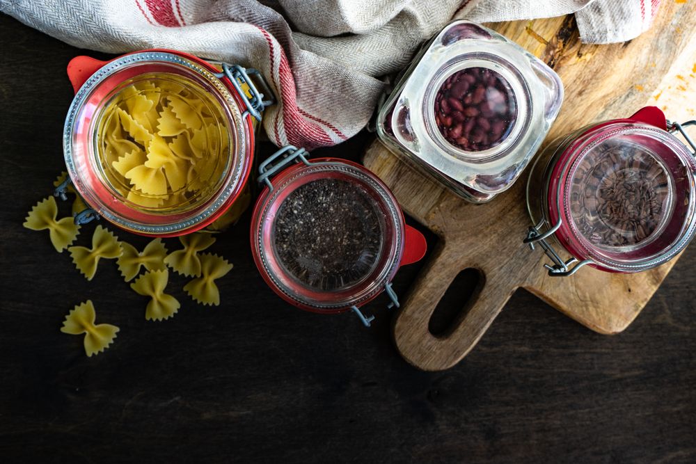Top view of ingredients in glass containers with farfelle pasta, seeds and beans on wooden background with copy space