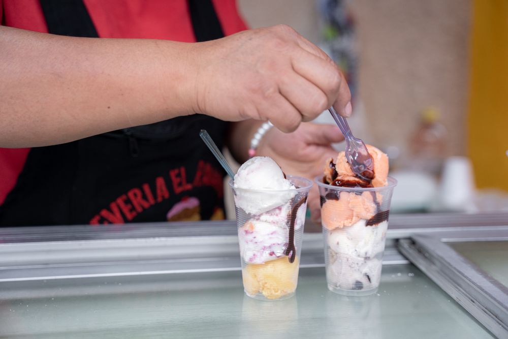 An Hispanic adult woman is serving some ice cream balls with a spoon in plastic cups in the street