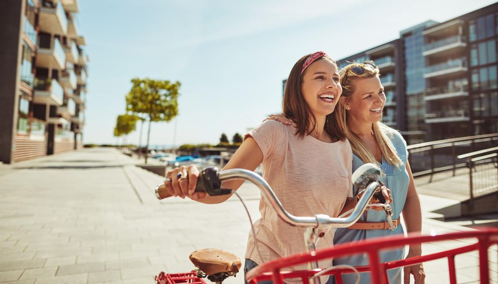 Two smiling female friends laughing while walking along a riverwalk with bike on sunny day