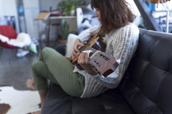 Woman sitting with crossed legs on sofa playing acoustic guitar