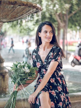 Charming female resting on edge of fountain with bouquet