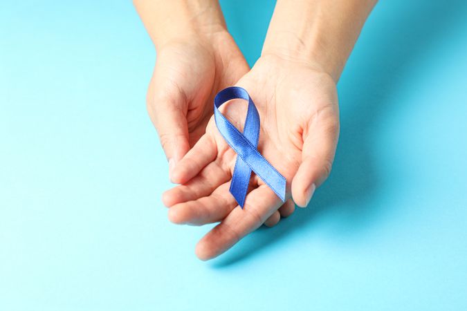 Female hands hold blue awareness ribbon on color background, space for text