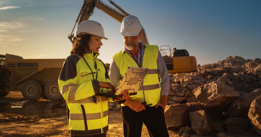 Male Engineer And Hispanic Female Building Architect Walking On Construction Site With Laptop Computer And Talking About New Real Estate Project. Heavy Machinery Working. Golden Hour