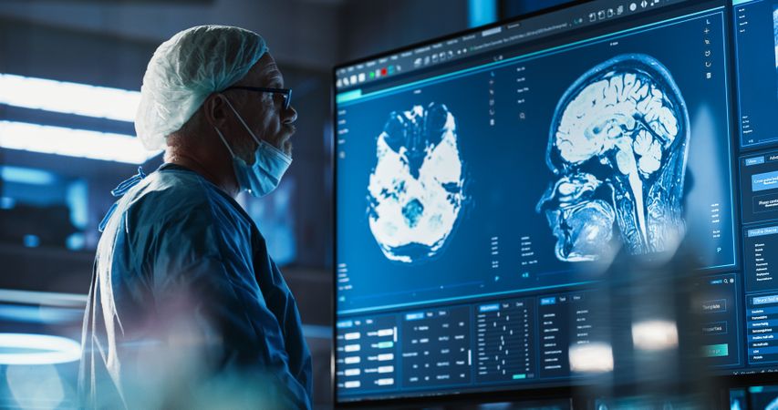 White Male Neurosurgeon Looking At TV Screen With Brain MRI Scans Of Patient, Analyzing, Preparing For Complicated Neurosurgery, Wearing Surgical Gown.
