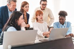 In a modern office setting, a redhead woman, sitting and leading a presentation on her laptop to diverse colleagues