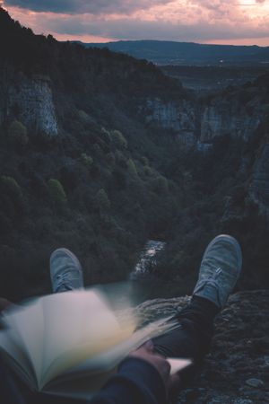 View to small river in canyon and legs of person with book sitting on the edge