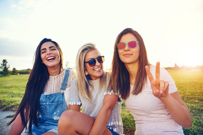 Laughing multi-ethnic female friends sitting in sunny field with one making the peace sign with hand