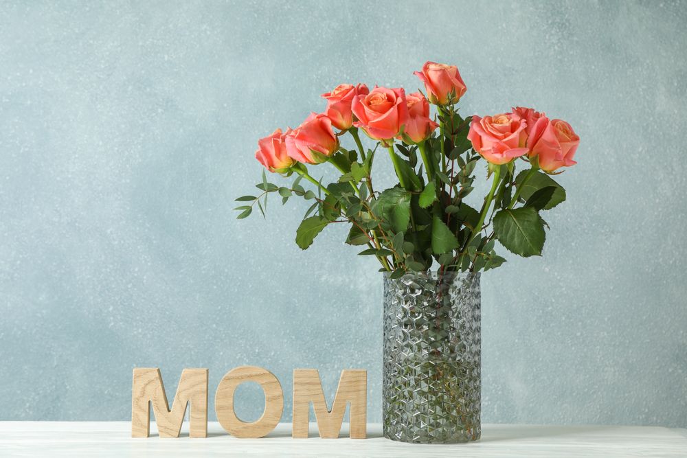 Vase with orange roses and inscription Mom on table against light background, space for text