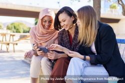 Three smiling women sitting on outdoor park bench talking while watching smartphone 5lzJeb