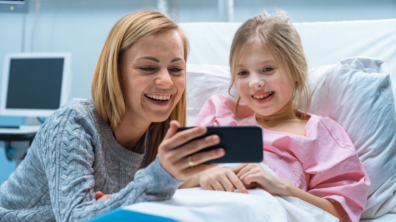 Cute Little Girl Lies on a Bed in the Children's Hospital, Her Mother Sits Beside, They Watch Cartoons/ Funny Videos on Smartphone