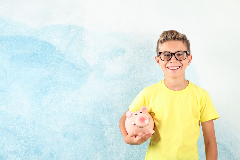 Boy in glasses with piggy bank against color background, space for text