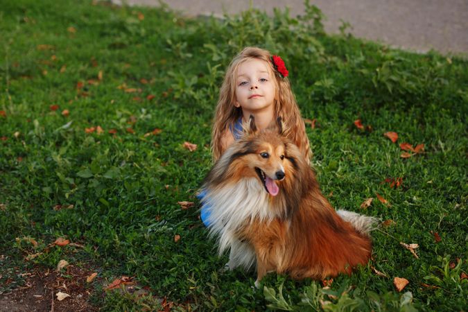 Female child in blue dress with flower in hair sitting with dog in the grass
