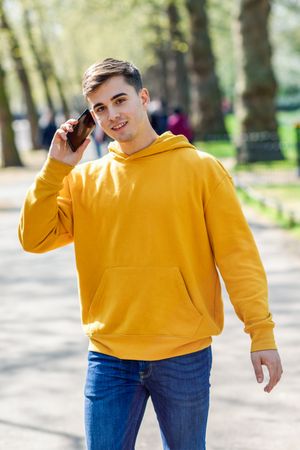 Man in yellow hoodie talking on cell phone while walking in park, vertical