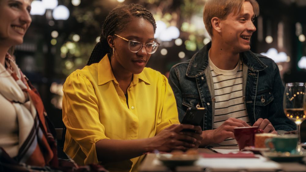 Young Black Female Focused On Smartphone, Not Paying Attention to a Group of Friends Sitting in a Cafe