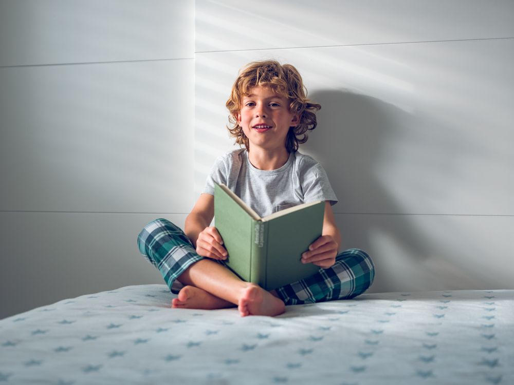 Cheerful curly blond boy reading book while sitting on bed